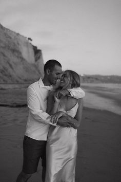 a man and woman standing next to each other on a beach near the ocean with cliffs in the background