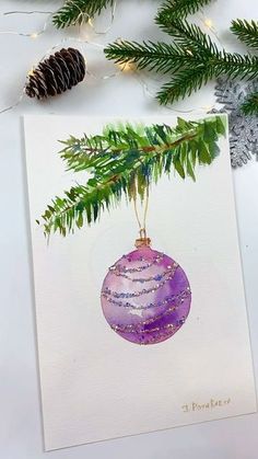 a watercolor christmas ornament hanging from a pine tree