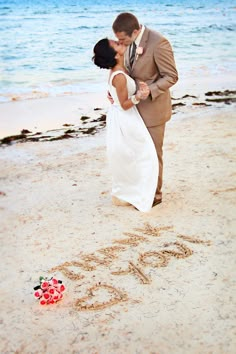 a bride and groom kissing on the beach in front of the word love written in the sand