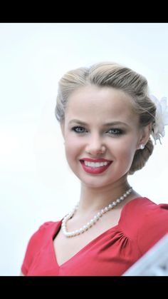 a woman in a red dress with pearls and a flower in her hair smiling at the camera