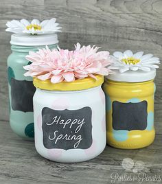 three painted mason jars with flowers in them sitting on a wooden table, one has a chalkboard sign and the other says happy spring