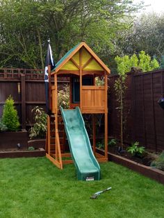 a wooden play set with a green slide in the grass next to a brown fence