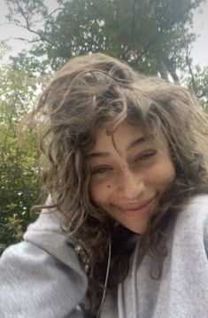 a woman with curly hair is taking a selfie in front of some trees and bushes