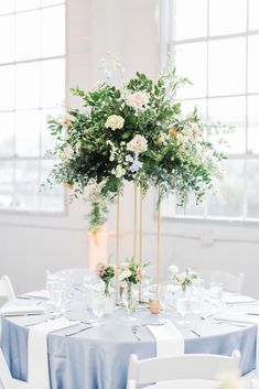 a tall vase filled with white flowers and greenery on top of a blue table cloth