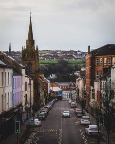 a city street with cars parked on both sides and a church steeple in the background