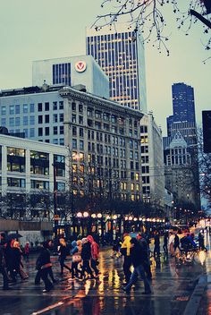 many people are crossing the street in the city on a rainy day with buildings behind them