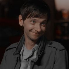 a young man is smiling and looking at the camera while wearing a gray jacket with an open collar