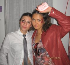 two women are posing for the camera in front of a curtained window, one is wearing a red leather jacket and the other wears a white shirt