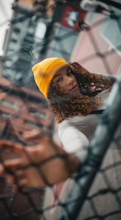 a woman with curly hair wearing a yellow hat and looking through a chain link fence