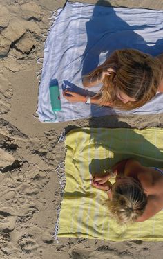 two women laying on towels in the sand with one holding a bottle and another looking down at her cell phone