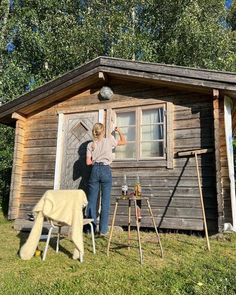 a woman standing in front of a small wooden cabin next to an easel and ladder