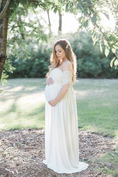 a pregnant woman in a white dress standing under a tree with her hands on her belly