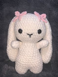 a crocheted white stuffed animal with pink bows on its head and ears, standing in front of a black background