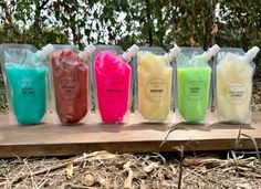 five different colored soaps are lined up on a wooden board in front of some trees