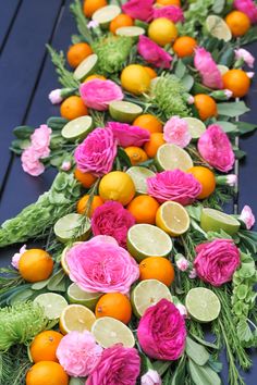 an arrangement of flowers and fruit arranged on a long table with dark blue boards in the background