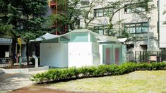 a small white building sitting in the middle of a lush green park next to tall buildings