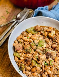 a white bowl filled with beans and meat on top of a wooden table next to silverware