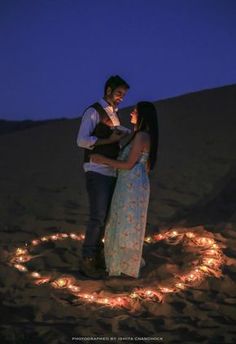 a man and woman standing in the sand holding each other with lights on their backs