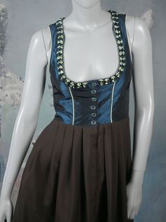 This German dirndl dress has a peacock blue satin bodice with a deep scoop neckline edged in a blue and mint green Tudor ruffle (the same green is used on the contouring lines on the front and back of the bodice). The dress closes in the front with six aquamarine rhinestone buttons to the waistline and two concealed buttons on the skirt. The dress has a fitted waistline that gives way to a softly pleated above-the-knee skirt (with a concealed pocket on the right side). The top is lined in a dark Fitted Blue Dress For Oktoberfest, Blue Fitted Dress For Oktoberfest, German Dirndl Dress, German Dress Dirndl, Silver Evening Dress, Knee Skirt, Dirndl Dress, Knee Skirts, 1960s Fashion