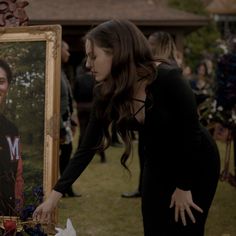 a woman in a black dress holding a framed photo next to a painting on the grass