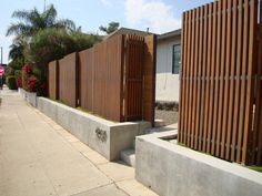 a wooden fence next to a sidewalk with flowers on the side and another building in the background