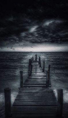 a pier in the ocean with birds flying over it and dark clouds above, on a cloudy day