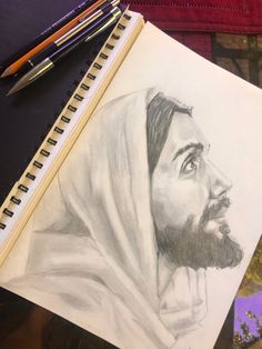 a pencil drawing of jesus on paper next to two pens and a notebook with the image of jesus