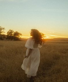 a woman in a white dress is walking through the grass at sunset with her hair blowing in the wind