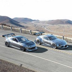 two silver sports cars driving down the road next to each other in front of mountains