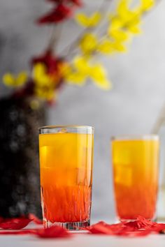 two glasses filled with liquid sitting on top of a table next to red and yellow flowers