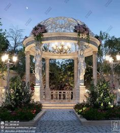 an outdoor gazebo surrounded by flowers and greenery at dusk with lanterns on the ceiling