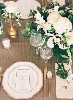 the table is set with white flowers and greenery, gold cutlery, and silverware