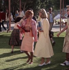 three women in dresses are talking to each other on the grass near people walking around