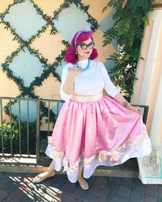 a woman with pink hair and glasses is posing on a bench wearing a white shirt and pink skirt