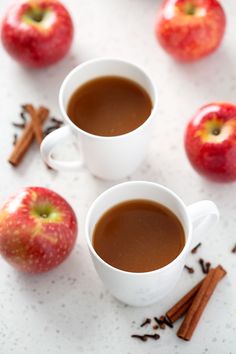 two cups of hot chocolate next to cinnamon sticks and apples on a white countertop