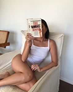 a woman sitting in a chair reading a book with her legs crossed on the pillow