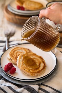 pancakes with syrup being poured onto them and raspberries on the plate next to them