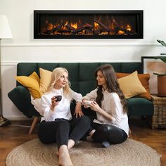 two women sitting on the floor in front of a fire place holding coffee cups and looking at their cell phones