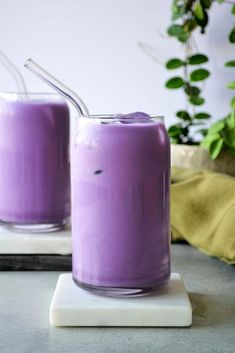 two glasses filled with purple liquid sitting on top of a white plate next to a green plant