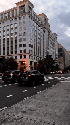 several cars are driving down the street in front of tall buildings at dusk, with traffic lights on