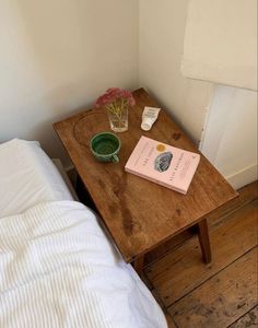 a small wooden table with a book and flower in a vase on it next to a bed