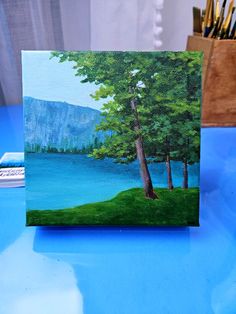 an acrylic painting of a lake and trees on a blue table with paintbrushes