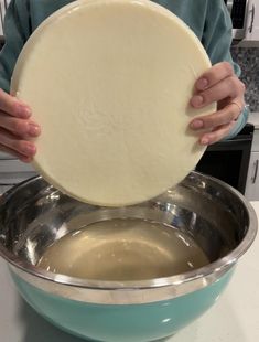 a person holding a pizza dough in a bowl on top of a counter with a metal pan