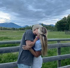 a man and woman kissing in front of a wooden fence with mountains in the background