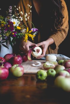 a woman is peeling an apple on a cutting board surrounded by apples and flowers in the background