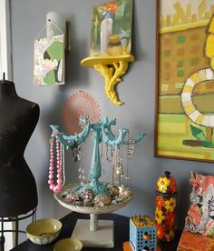 a table topped with lots of jewelry next to a wall mounted art piece on a gray wall