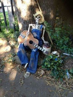 a skeleton sitting on the ground with a guitar in his lap next to a tree