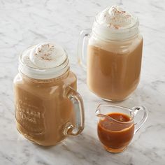 two mason jars filled with hot chocolate and topped with whipped cream, sit on a marble surface