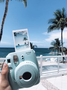 a person holding up a blue camera in front of the ocean with palm trees behind them