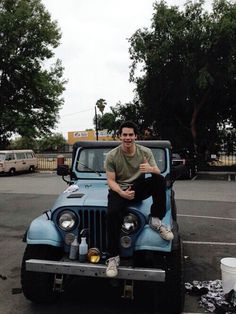 a man sitting on the hood of a blue jeep in a parking lot with trees